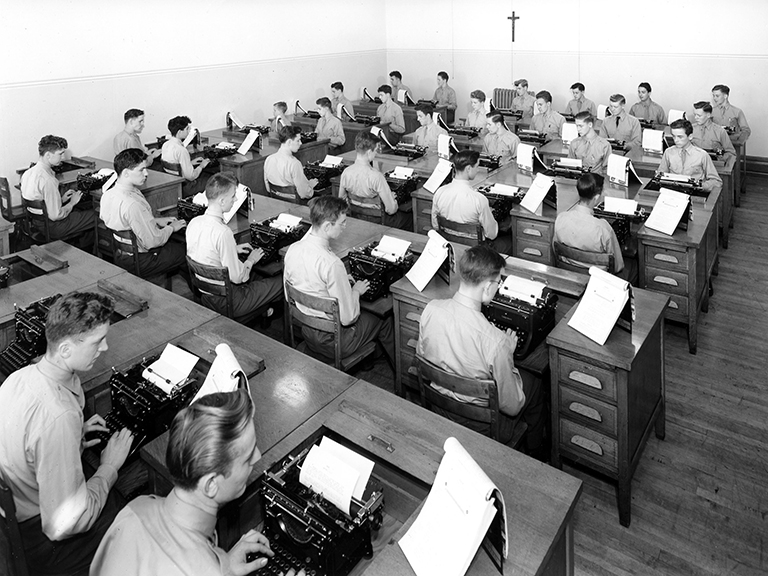 Dozens of young boys, well-dressed in button-down shirts, ties and slacks, sit at desks in rows and type on typewriters.  Half of the rows and students face the camera and half of the rows have their back to the camera. 